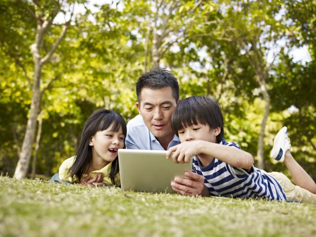 A man with his daughter and son, laying on the grass and using a tablet